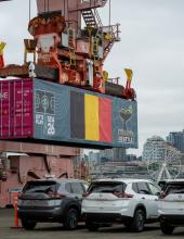 A shipping container with a Belgium flag banner hangs from a crane at Terminal 46 in Seattle.